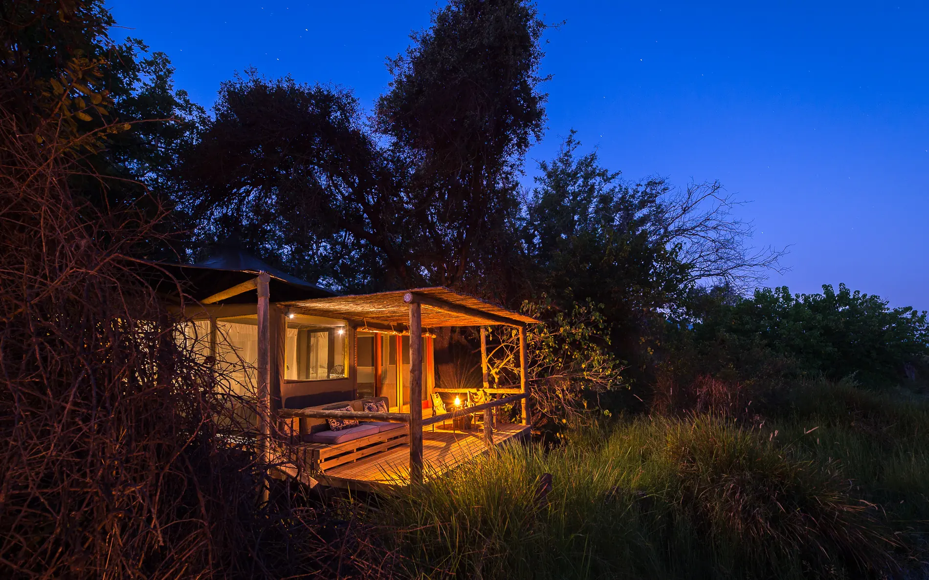 Illuminated safari lodge surrounded by trees and grassland at dusk, Little Vumbura, The Okavango Delta, Botswana