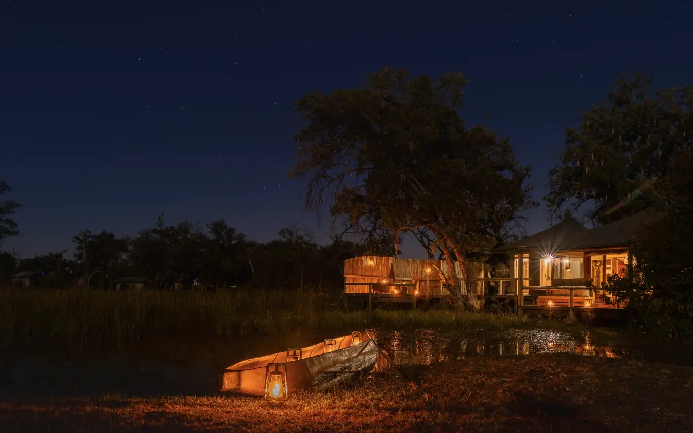 Illuminated safari lodge with canoe on riverbank at night under stars, Little Vumbura, The Okavango Delta, Botswana