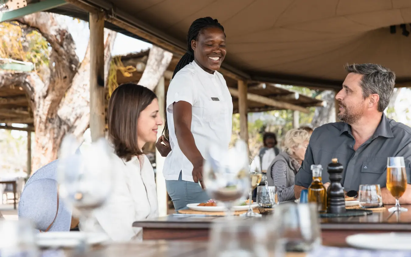 Server attending to guests at outdoor dining table under thatched shelter, Little Vumbura, The Okavango Delta, Botswana