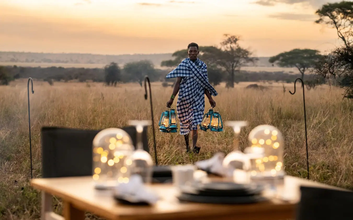 Staff member carrying lanterns toward outdoor dinner setup at sunset, Lolkisale Camp, Tarangire National Park, Tanzania