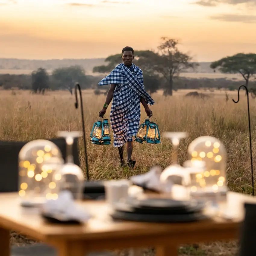 Staff member carrying lanterns toward outdoor dinner setup at sunset, Lolkisale Camp, Tarangire National Park, Tanzania