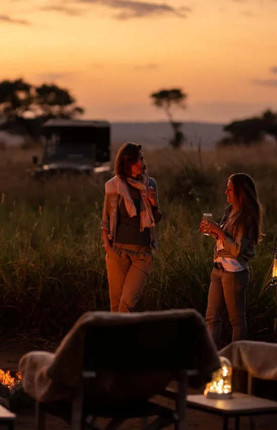 Two guests standing by campfire at sunset in savanna, Lolkisale Camp, Tarangire National Park, Tanzania