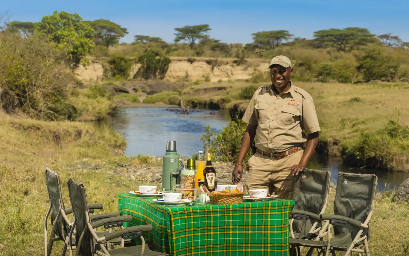 Safari guide standing beside outdoor dining setup near river, Mahali Mzuri, Maasai Mara, Kenya