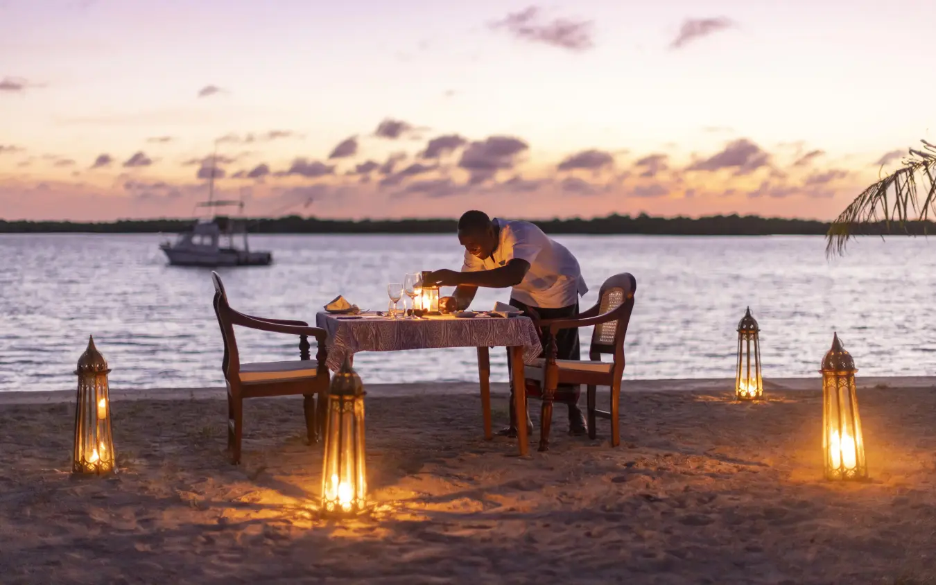 Beach dinner setup at sunset with server and sailboat, Manda Bay, Lamu Archipelago, Kenya