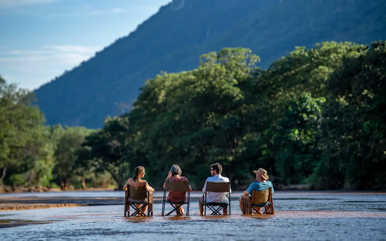 Four guests sitting in chairs in shallow river water, Manyara Green Camp, Lake Manyara, Tanzania