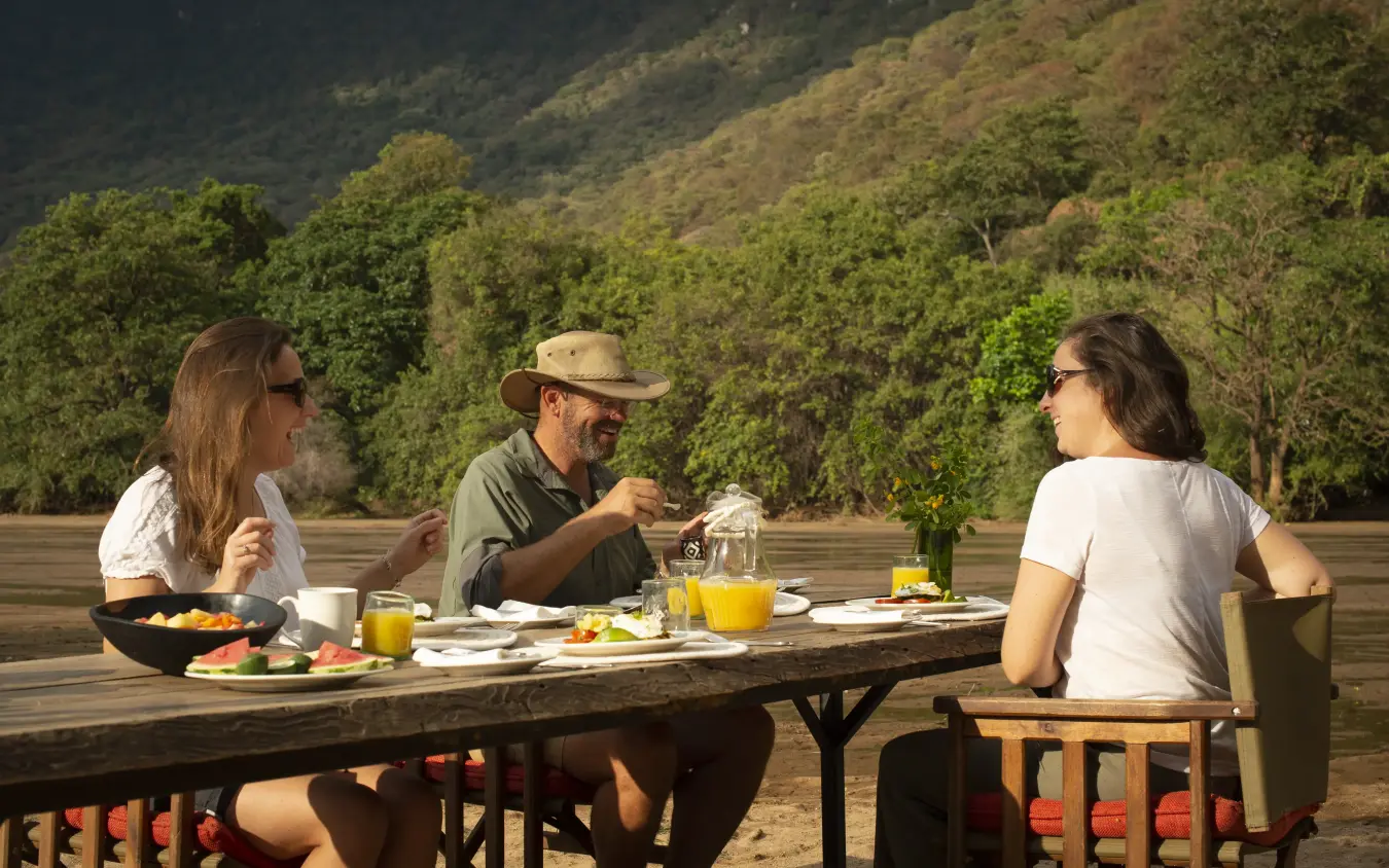 Guests dining outdoors on wooden deck overlooking forested river valley, Manyara Green Camp, Lake Manyara, Tanzania