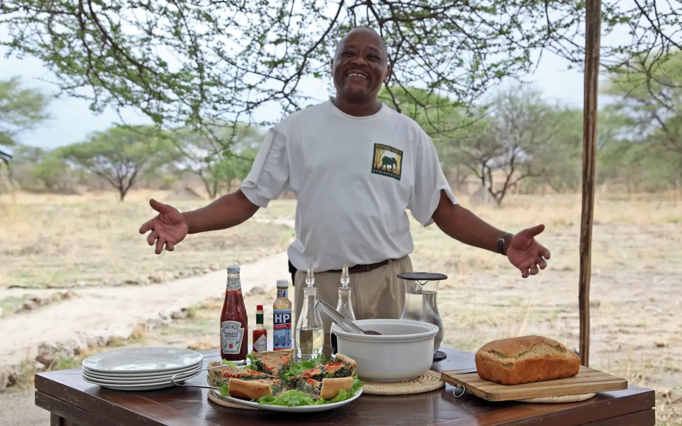 Chef presenting outdoor meal setup under acacia trees in savanna landscape, Manyara Ranch, Lake Manyara, Tanzania