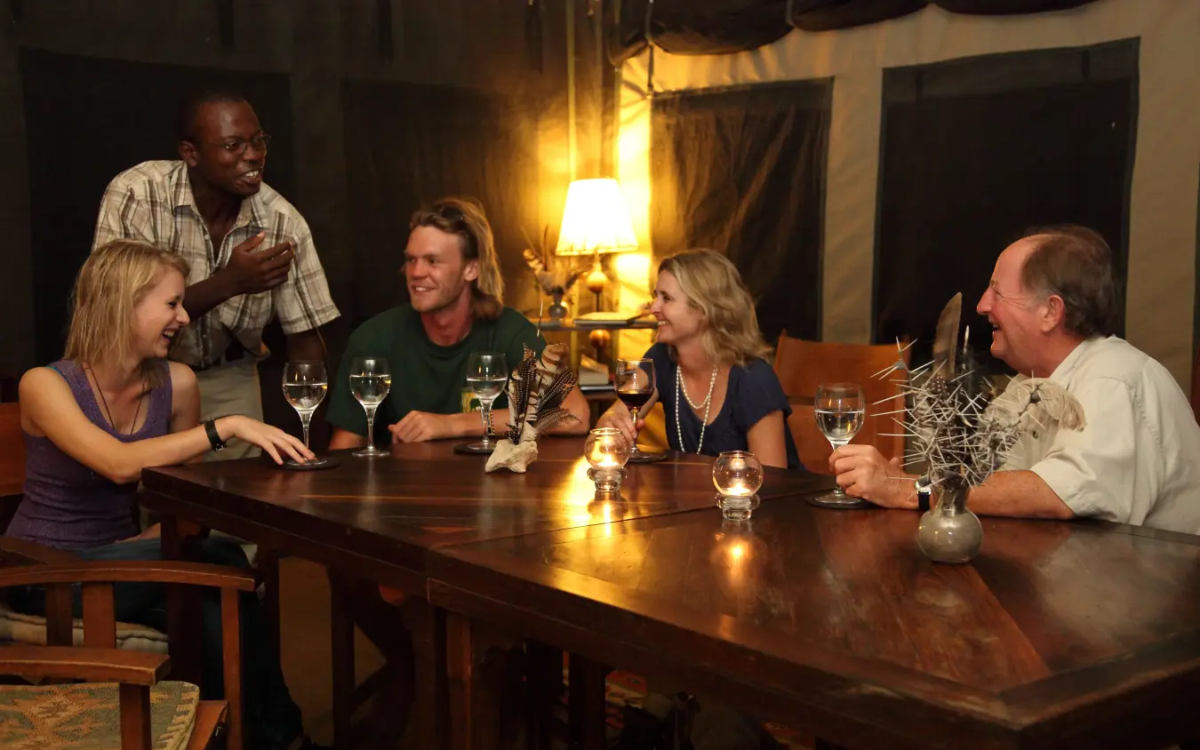 Group of guests dining together at wooden table in evening light, Manyara Ranch, Lake Manyara, Tanzania