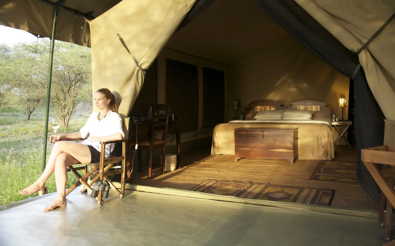 Guest relaxing in canvas safari tent with wooden bed and savanna view, Manyara Ranch, Lake Manyara, Tanzania