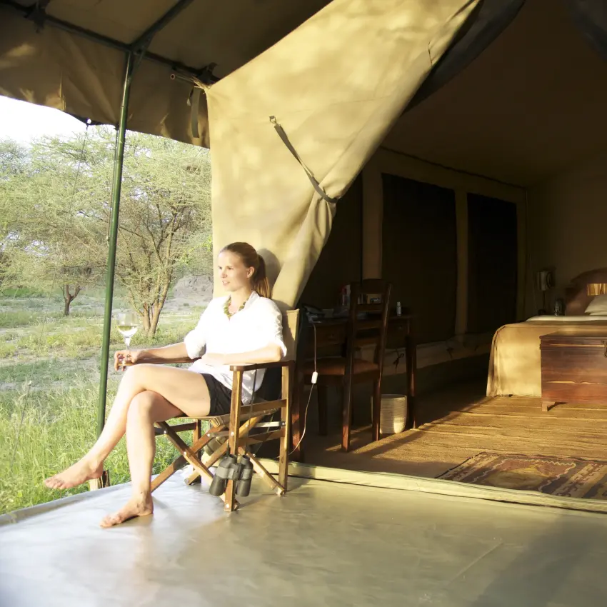 Guest relaxing in canvas safari tent with wooden bed and savanna view, Manyara Ranch, Lake Manyara, Tanzania