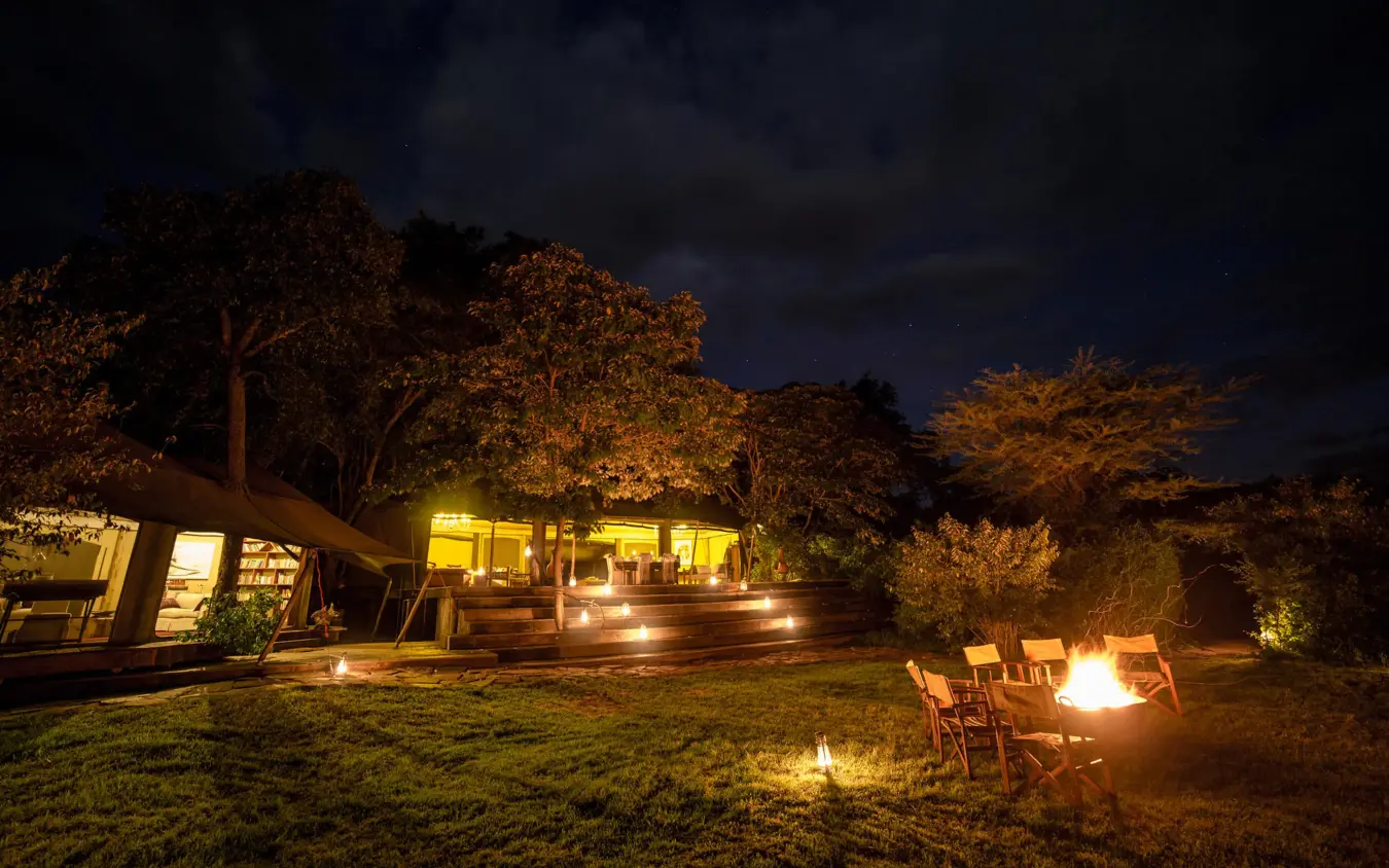 Illuminated lodge buildings with fire pit seating area at dusk, Mara Plains Camp, Maasai Mara, Kenya