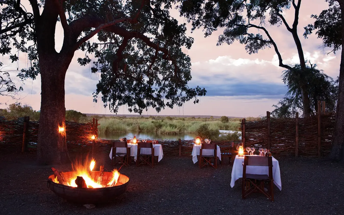 Outdoor dining setup by firelight under large trees, Matetsi River Lodge, Victoria Falls, Zimbabwe