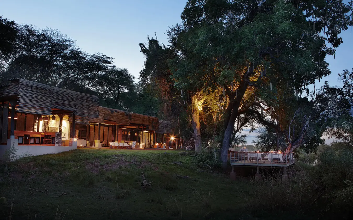 Illuminated lodge building nestled beneath large ancient tree at dusk, Matetsi River Lodge, Victoria Falls, Zimbabwe