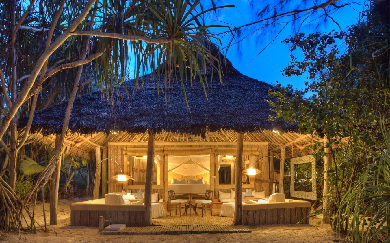 Beachfront thatched bungalow illuminated at dusk with palm trees, Mnemba Island, Zanzibar & Pemba, Tanzania