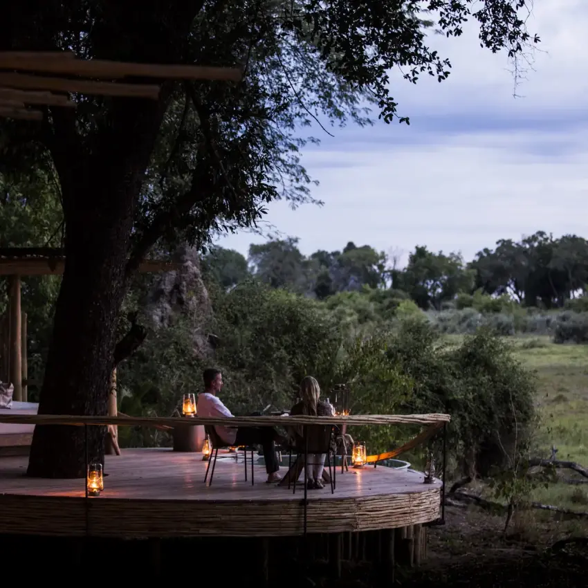 Wooden deck dining area beneath ancient tree overlooking savanna landscape at dusk, Mombo Camp, The Okavango Delta, Botswana