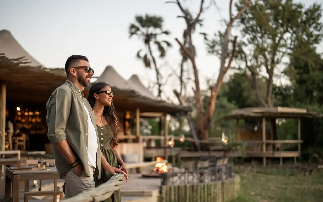 Couple at safari lodge viewing deck at dusk with acacia trees, Mombo Camp, The Okavango Delta, Botswana