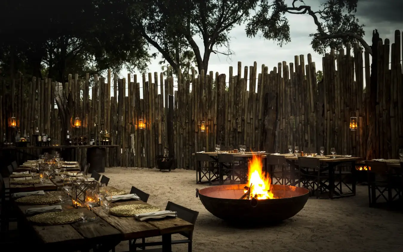 Outdoor dining area with central fire pit and wooden palisade fence at dusk, Mombo Camp, The Okavango Delta, Botswana