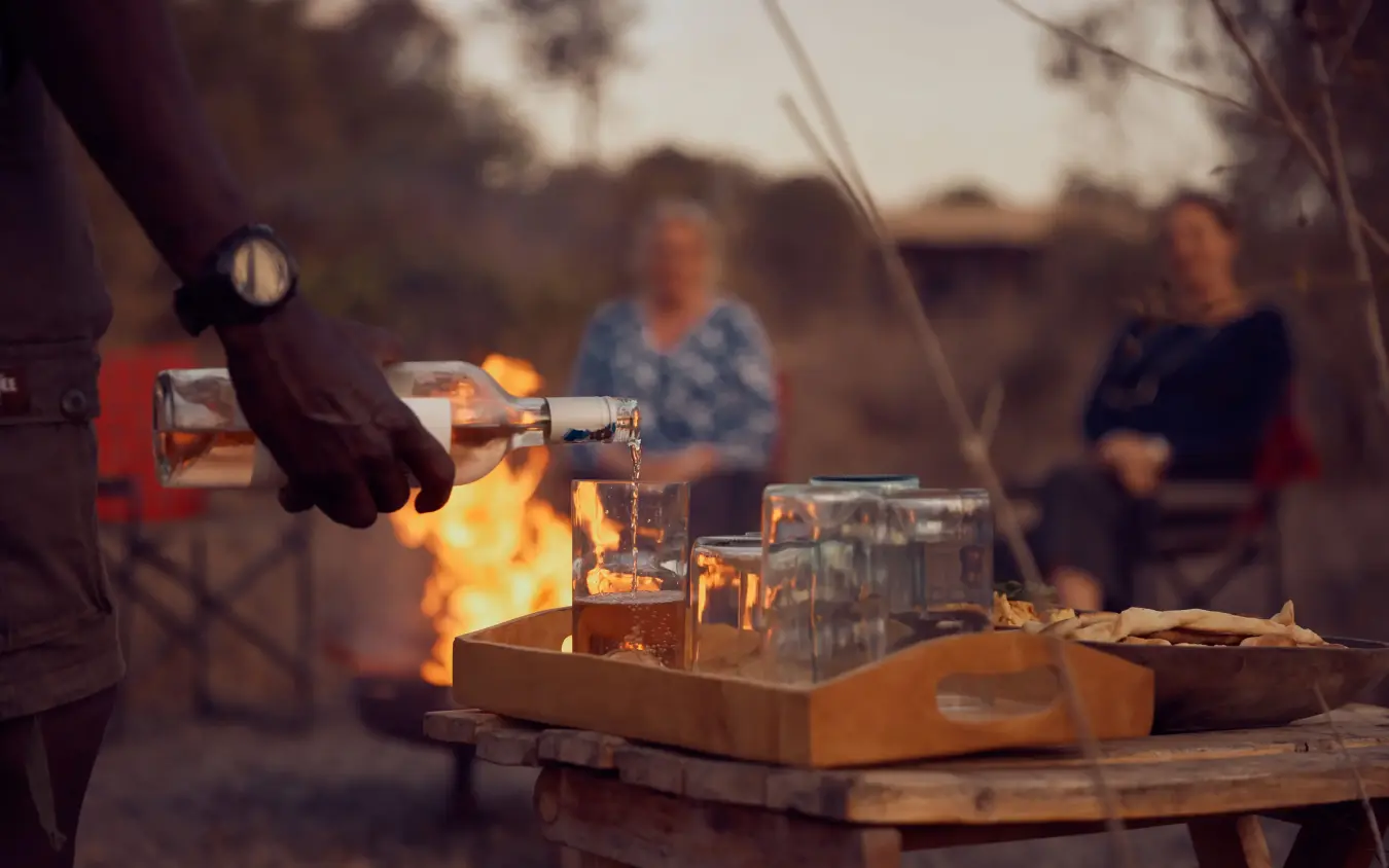 Person pouring drink by campfire with guests in background at dusk