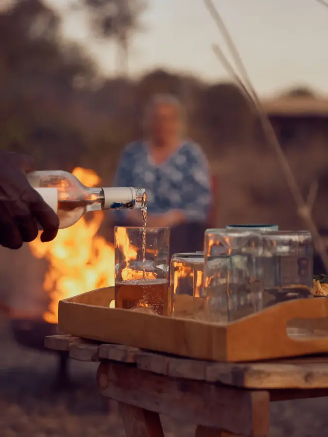 Person pouring drink by campfire with guests in background at dusk, Moon’s Camp, Laikipia Highlands, Kenya