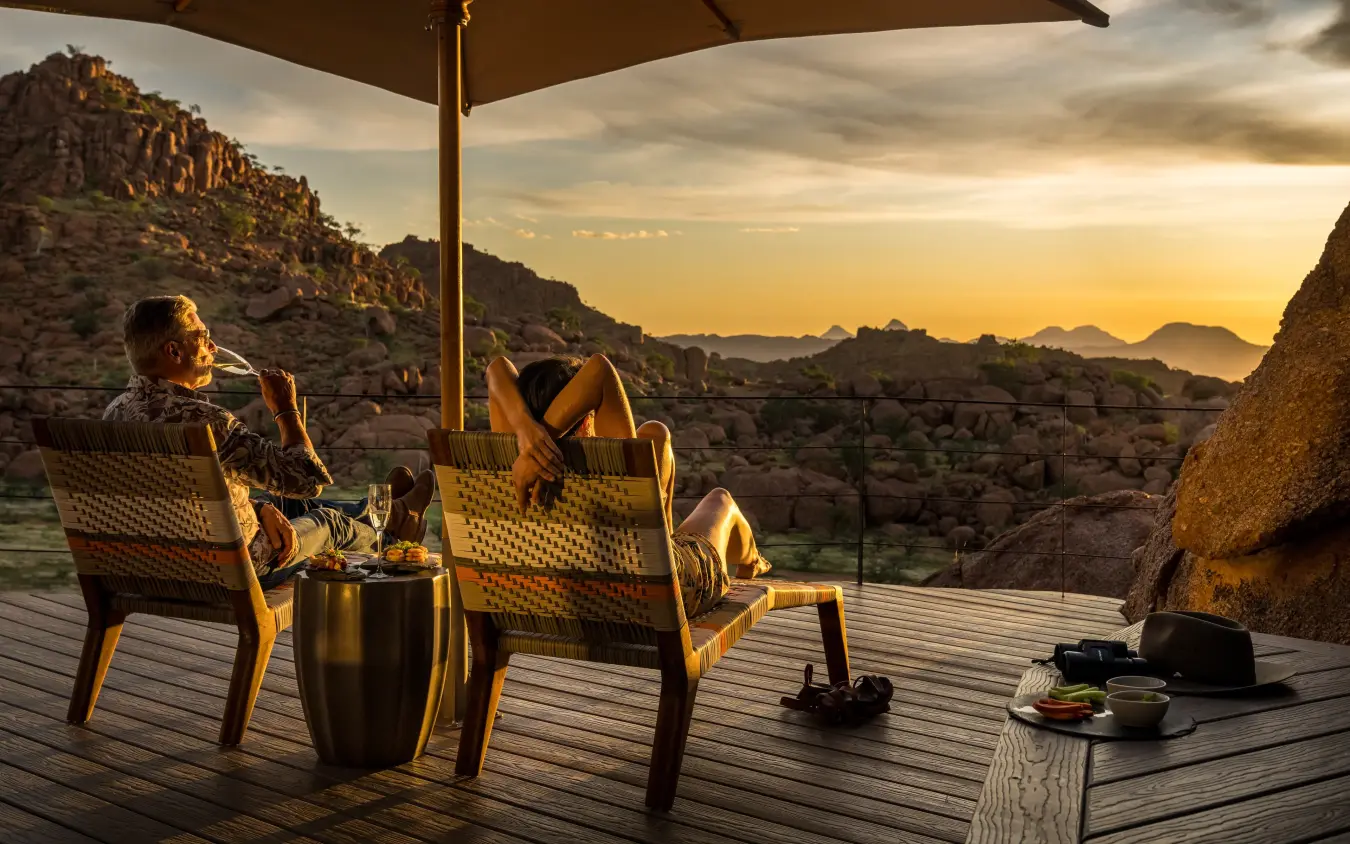 Couple relaxing on wooden deck at sunset with boulder rock formations, Mowani Mountain Camp, Damaraland, Namibia