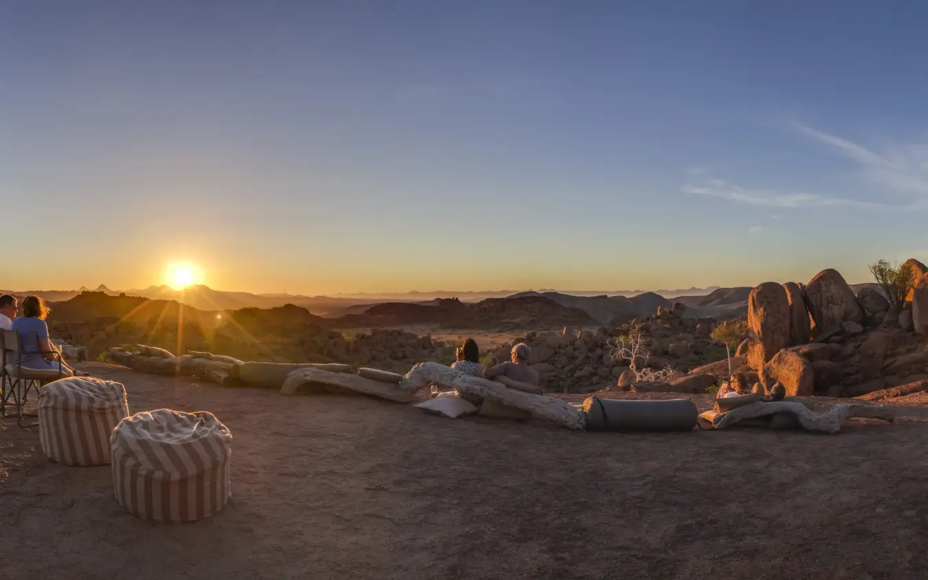 Guests watching sunset from rocky desert camp with log seating, Mowani Mountain Camp, Damaraland, Namibia