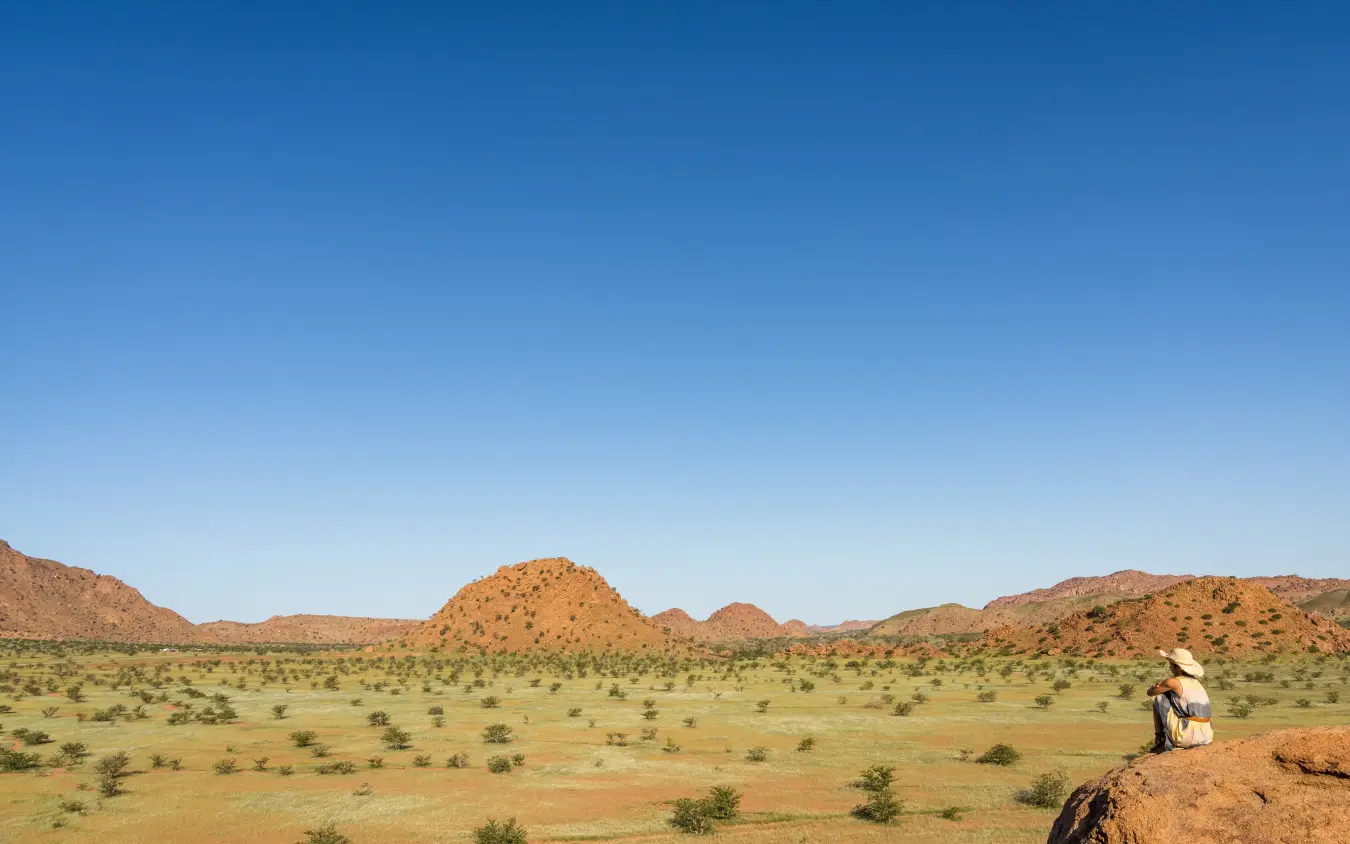Lone traveler sitting on rock overlooking arid grassland with granite hills, Mowani Mountain Camp, Damaraland, Namibia