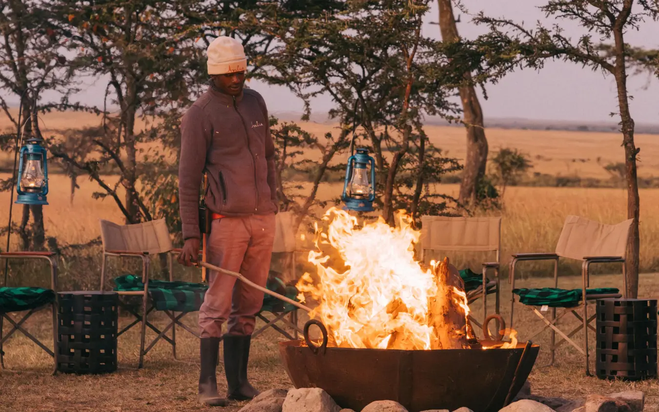 Guide tending campfire at evening gathering under acacia trees, Naboisho Camp, Maasai Mara, Kenya