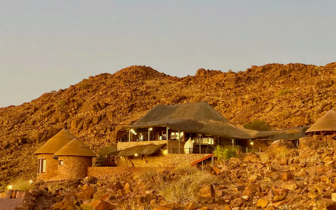 Luxury lodge with thatched roof nestled in rocky desert landscape at sunset, Namib Outpost, Sossusvlei, Namibia