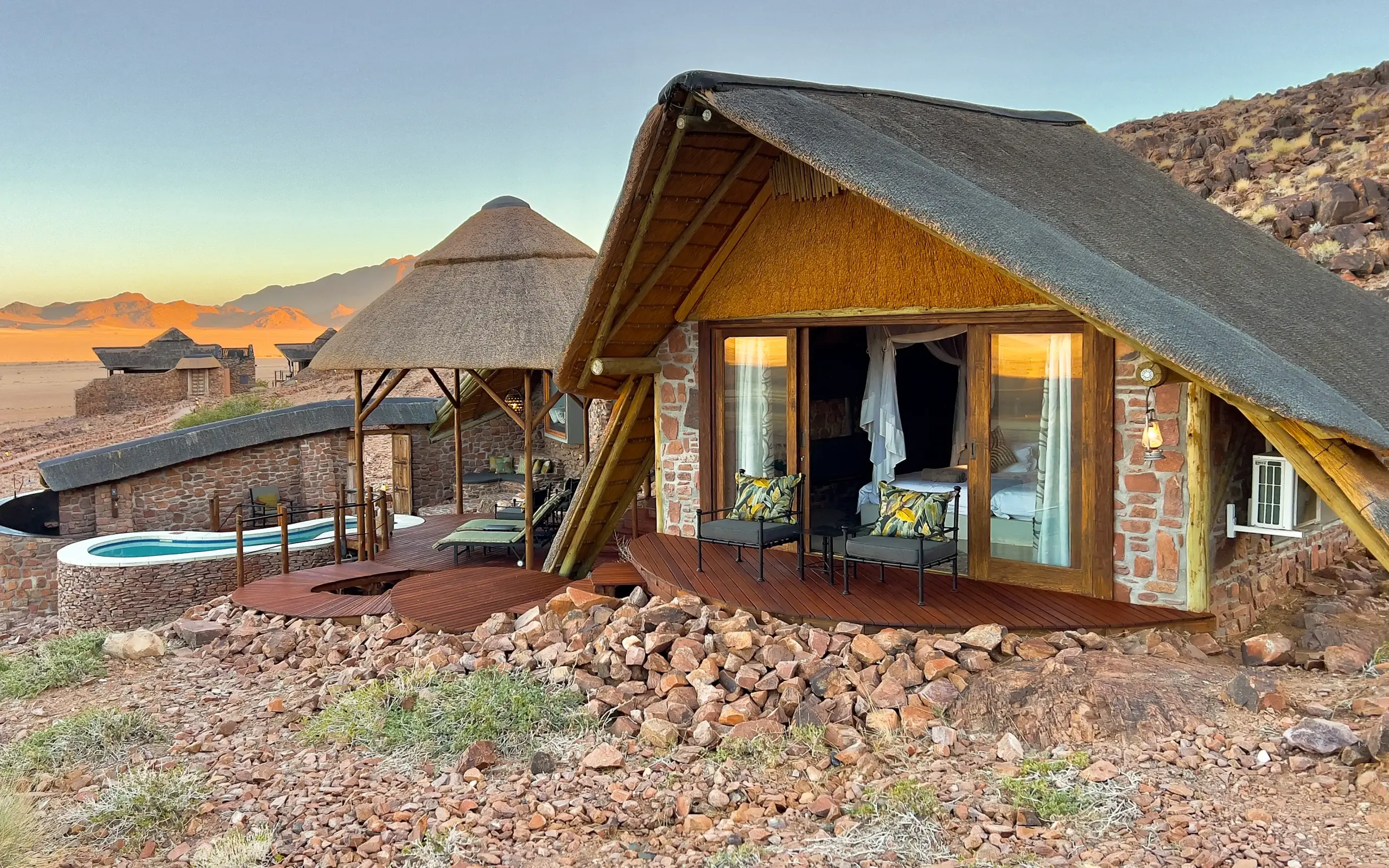 Luxury safari lodge with thatched roof overlooking desert mountain landscape at sunset, Namib Outpost, Sossusvlei, Namibia