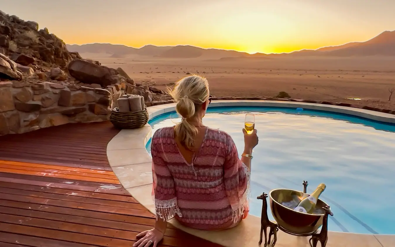 Woman with champagne relaxing by infinity pool overlooking desert landscape at sunset, Namib Outpost, Sossusvlei, Namibia