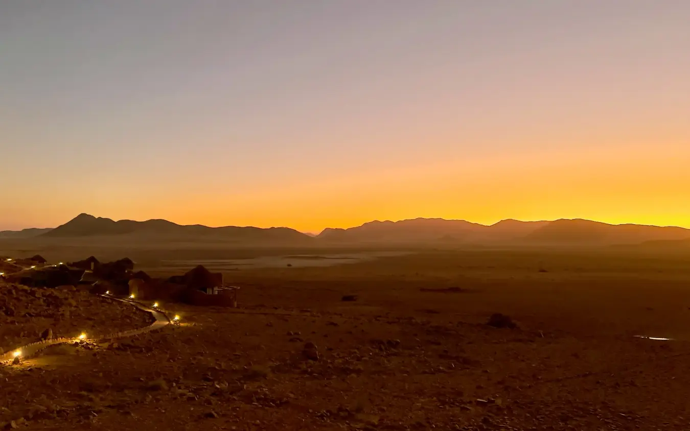 Desert landscape at dusk with illuminated safari lodge nestled in rocky terrain, Namib Outpost, Sossusvlei, Namibia
