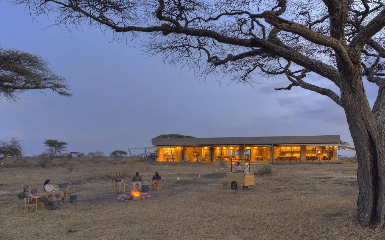 Illuminated lodge building at dusk with guests gathered around firepit, Namiri Plains, Serengeti National Park, Tanzania