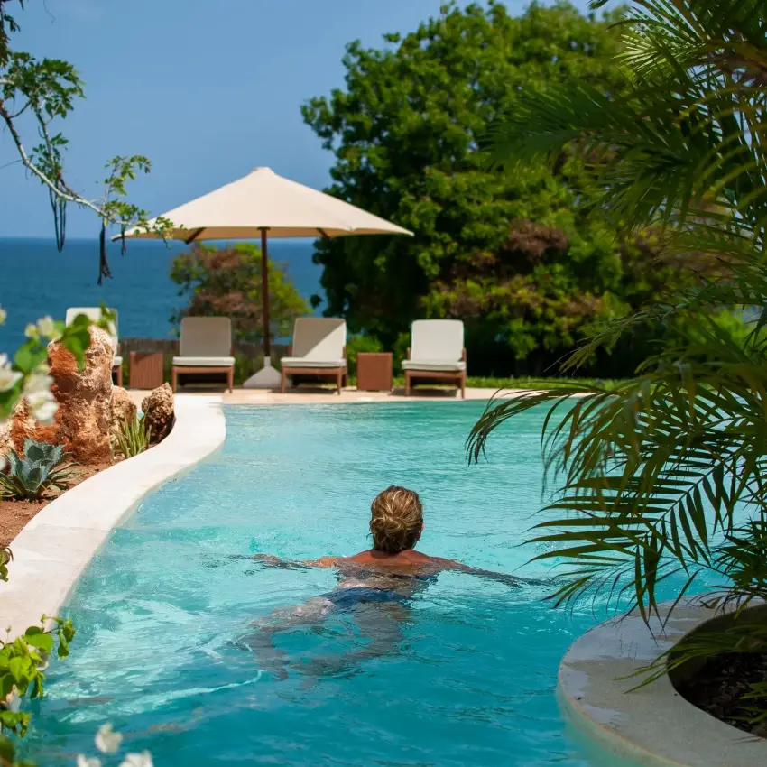 Guest swimming in infinity pool overlooking coastal ocean with loungers and umbrella, Nderit House, Kilifi, Kenya