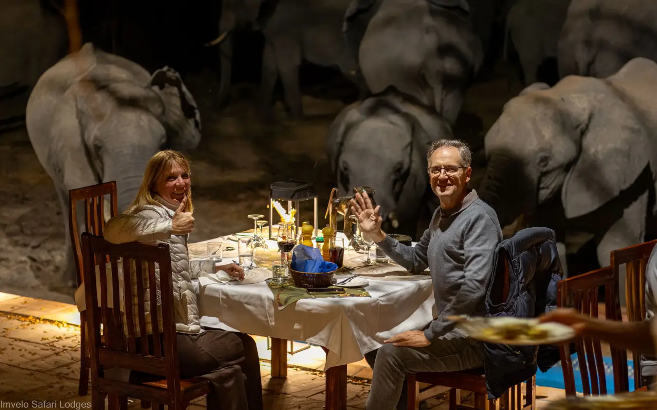 Couple dining at table with elephants in background at night, Nehimba Lodge, Hwange National Park, Zimbabwe