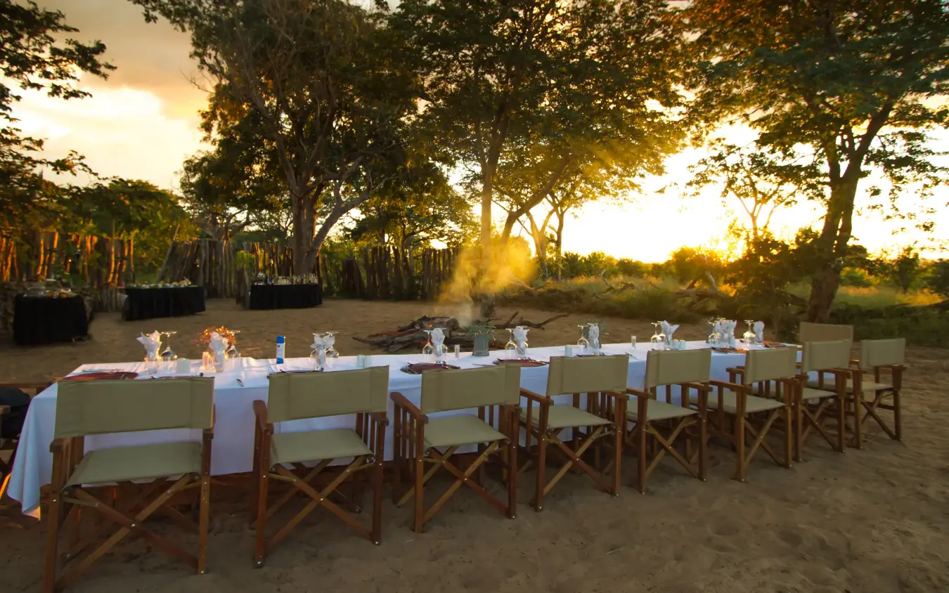 Outdoor dining setup on sandy ground at sunset under acacia trees, Ngoma Safari Lodge, Chobe, Botswana