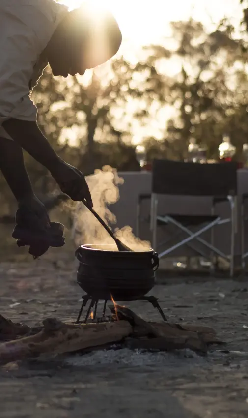 Person tending campfire at sunset during outdoor gathering, Kweene Trails, The Okavango Delta, Botswana