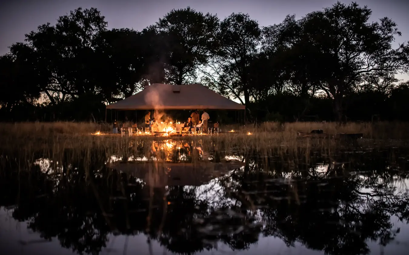 Illuminated safari lodge at dusk reflected in water, Kweene Trails, The Okavango Delta, Botswana