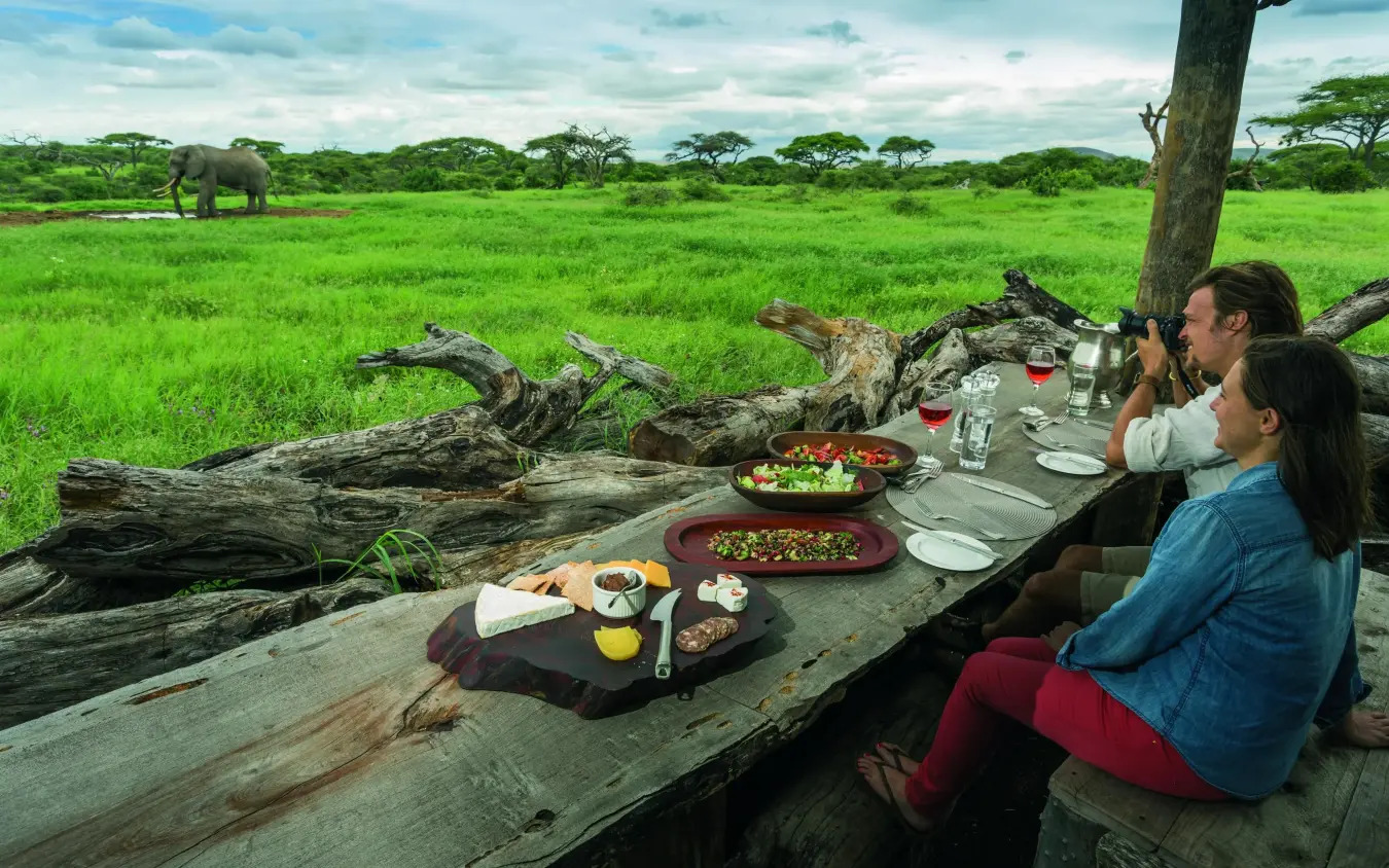 Guests dining outdoors with savanna landscape and wildlife, Ol Donyo Lodge, Amboseli and Chyulu Hills, Kenya