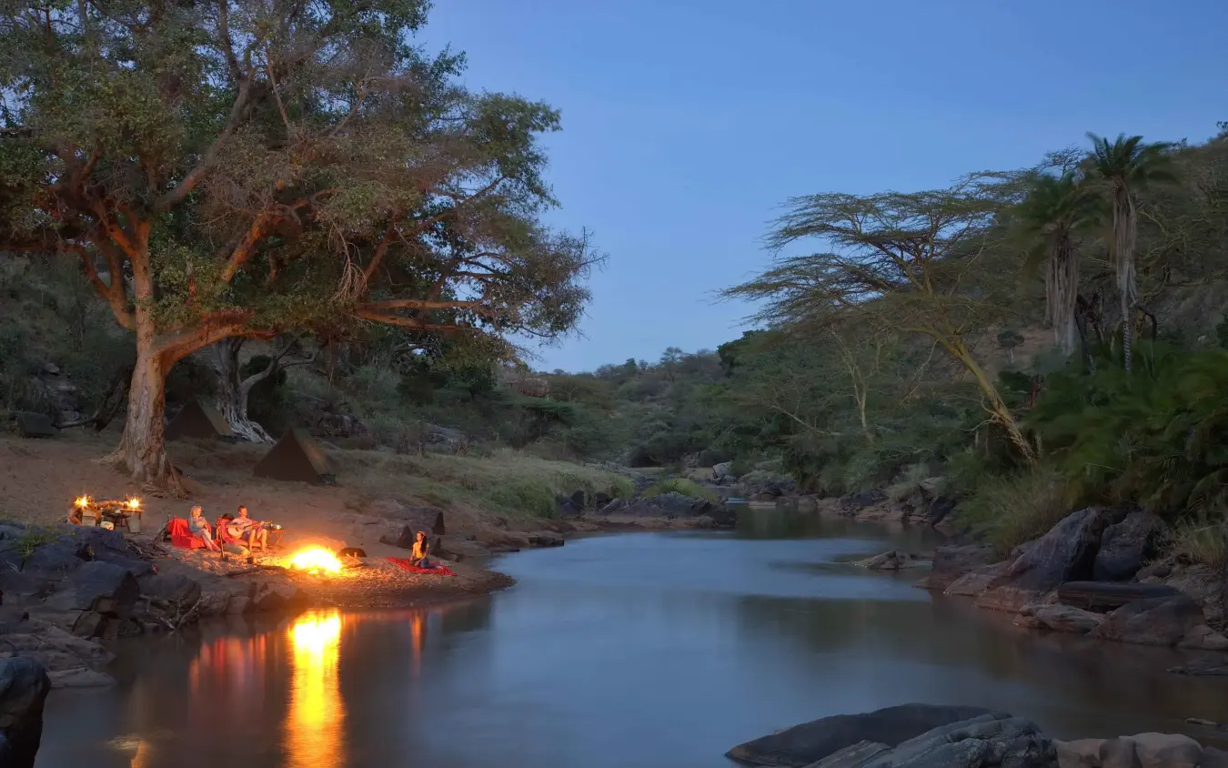 Guests gathered around campfire by river at dusk, Ol Malo Lodge, Laikipia Highlands, Kenya