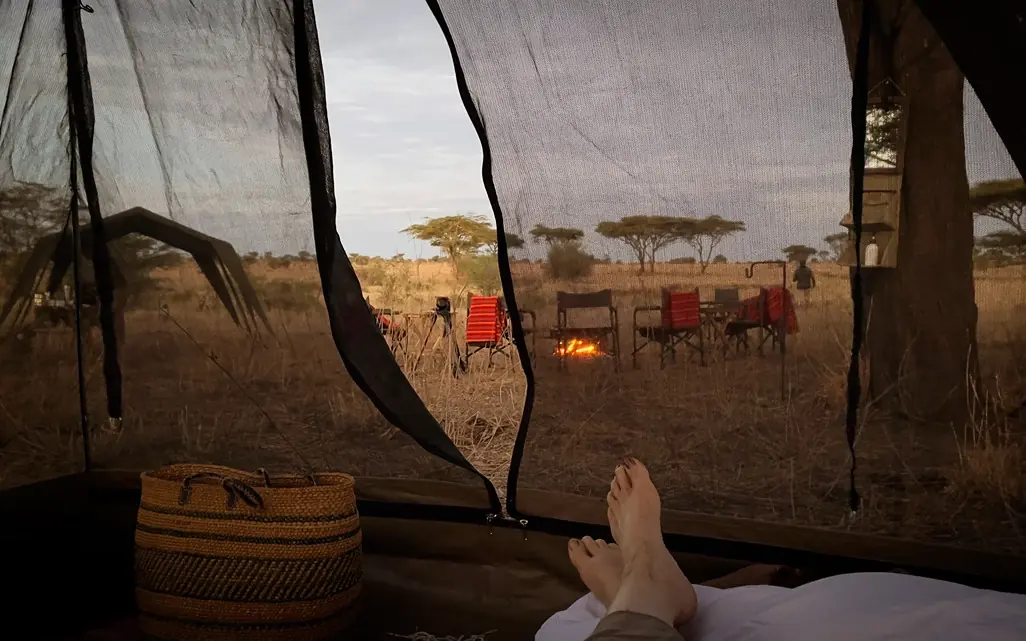 Bare feet relaxing in tent with savanna camp view through mesh, Olmara Serengeti, Serengeti National Park, Tanzania