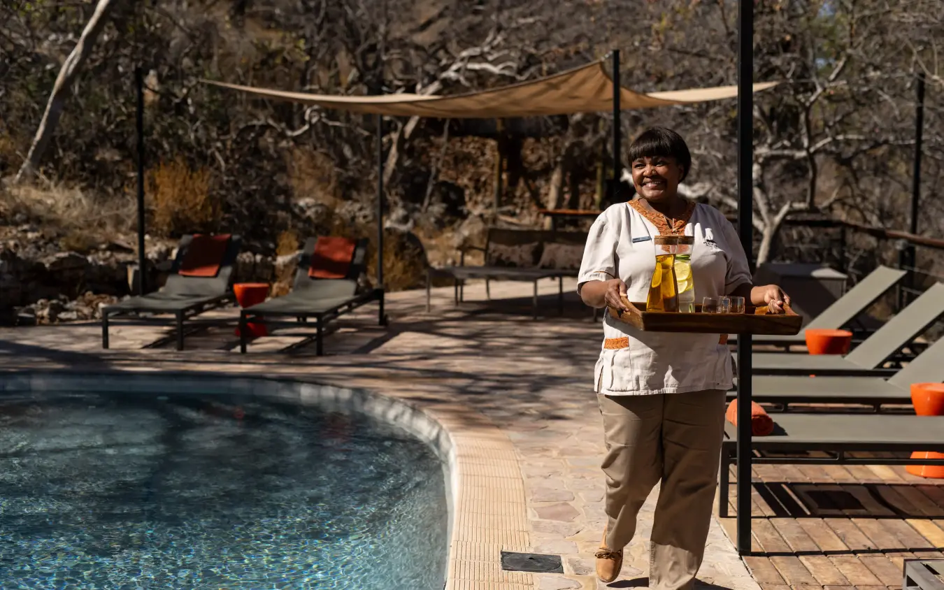 Staff member serving drinks by resort pool with loungers and shade structures, Ongava Lodge, Etosha National Park, Namibia
