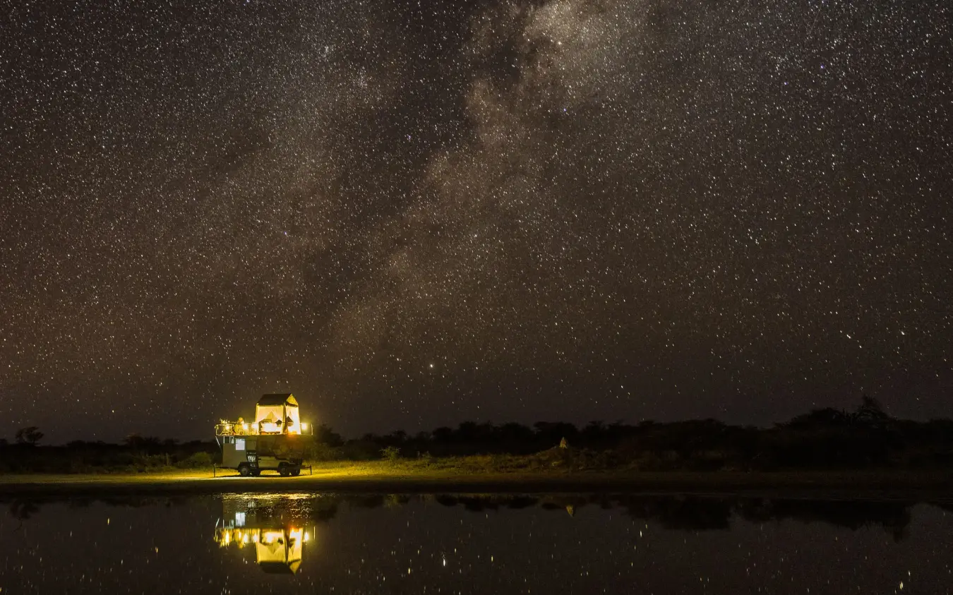 Illuminated safari lodge reflected in waterhole under starlit sky, Onguma Camp Kala, Etosha National Park, Namibia