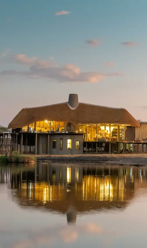 Luxury lodge with thatched roof reflected in waterhole at dusk, Onguma Camp Kala, Etosha National Park, Namibia