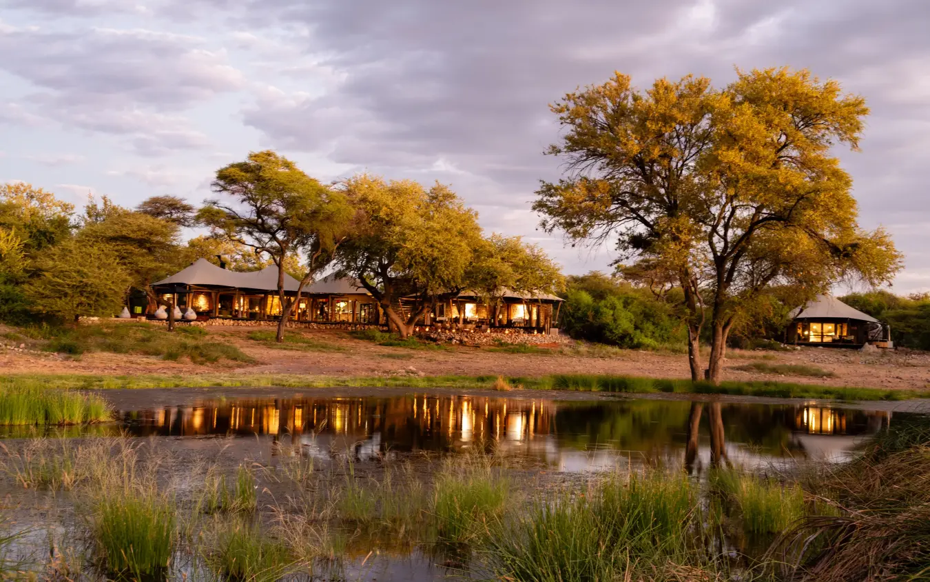 Riverside lodge with illuminated buildings reflected in waterhole at dusk, Onguma Tented Camp, Etosha National Park, Namibia