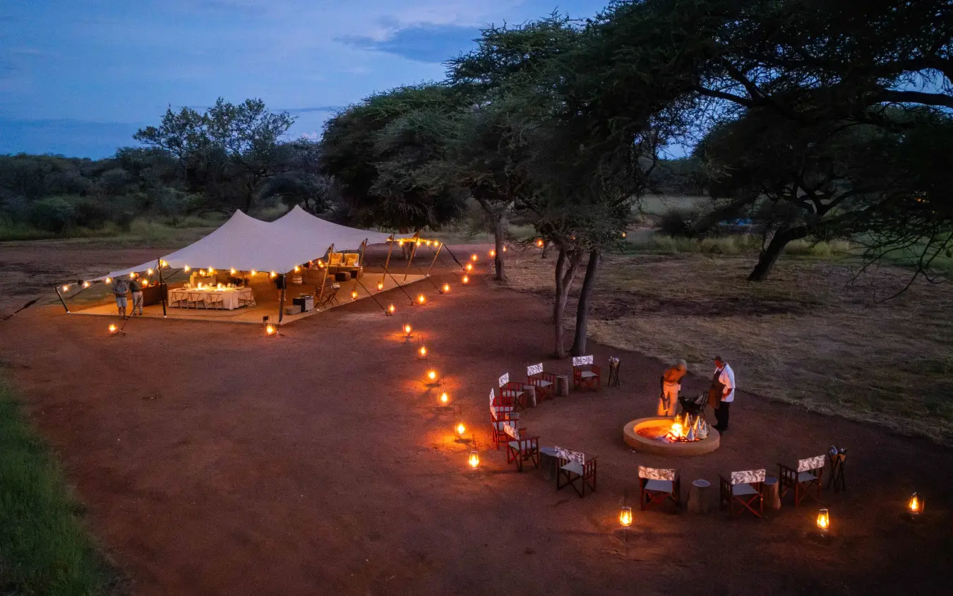 Candlelit outdoor dining setup with firepit under acacia trees at dusk, Onguma Trails Camp, Etosha National Park, Namibia
