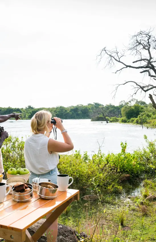 Guests observe wildlife with binoculars at riverside picnic setup, Papa’s Camp, Murchison Falls NP, Uganda