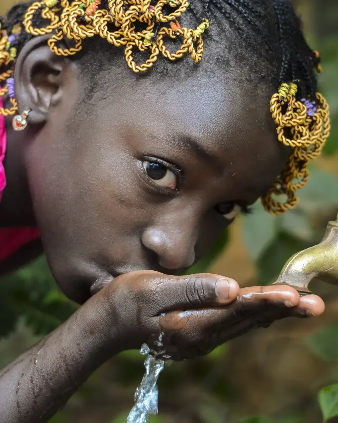Child drinking water from outdoor tap with green foliage