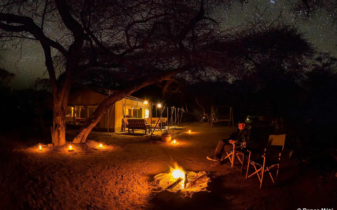 Glamping camp with firepit and illuminated tent under acacia tree at night, Porini Amboseli Camp, Amboseli and Chyulu Hills, Kenya