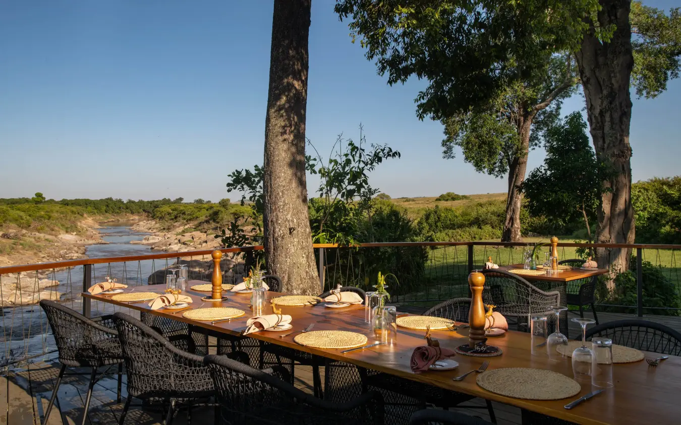 Outdoor dining setup under large trees overlooking river, Rekero Camp, Maasai Mara, Kenya