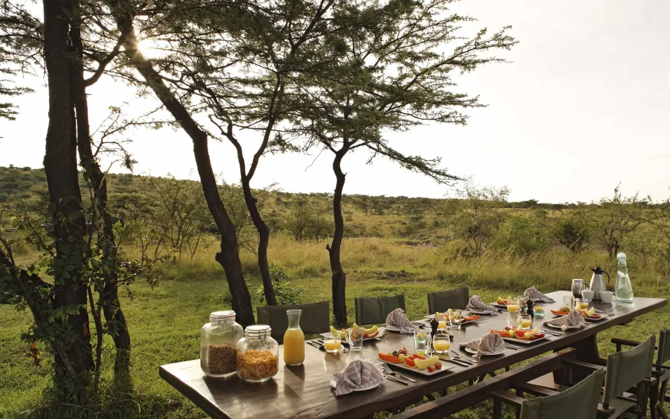 Outdoor dining table set under acacia trees with savanna landscape, Richard’s River Camp, Maasai Mara, Kenya